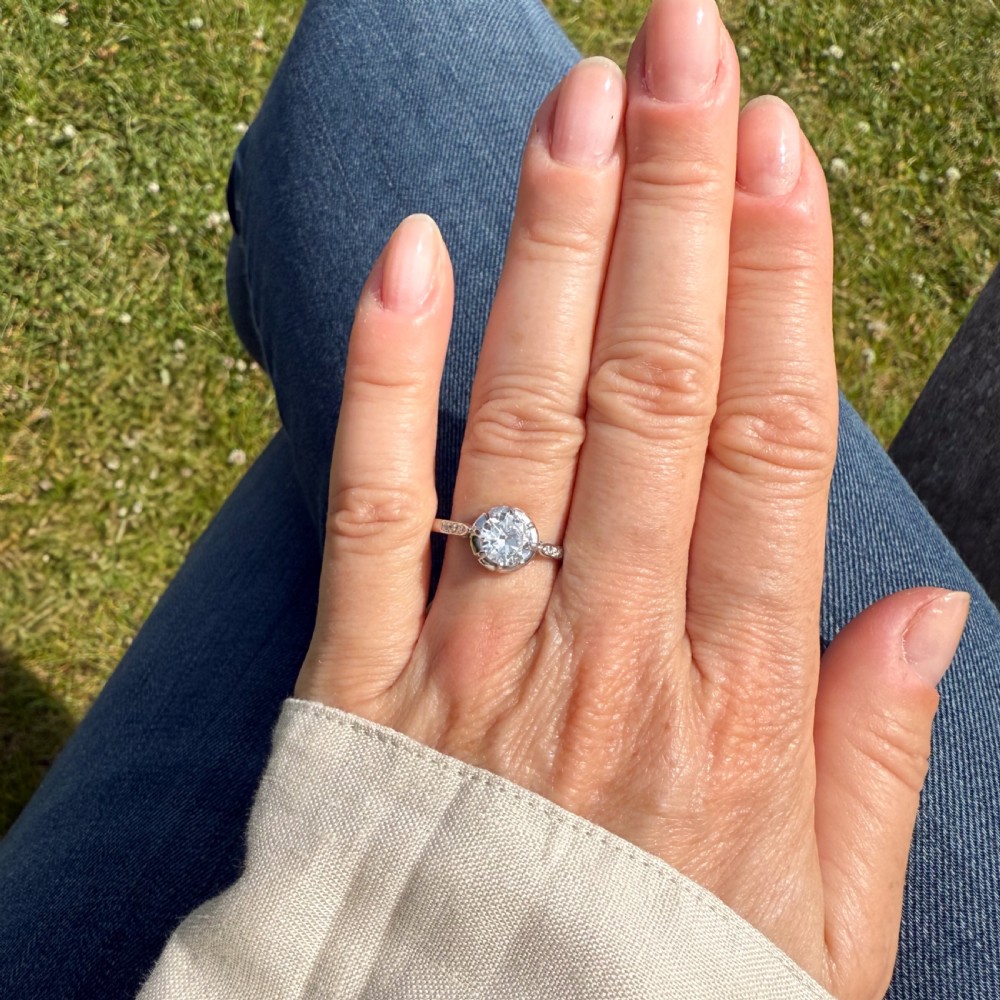 An Art Deco solitaire engagement ring featuring a central round old cut diamond set in white metal with accent diamonds set into the shoulders. View VI