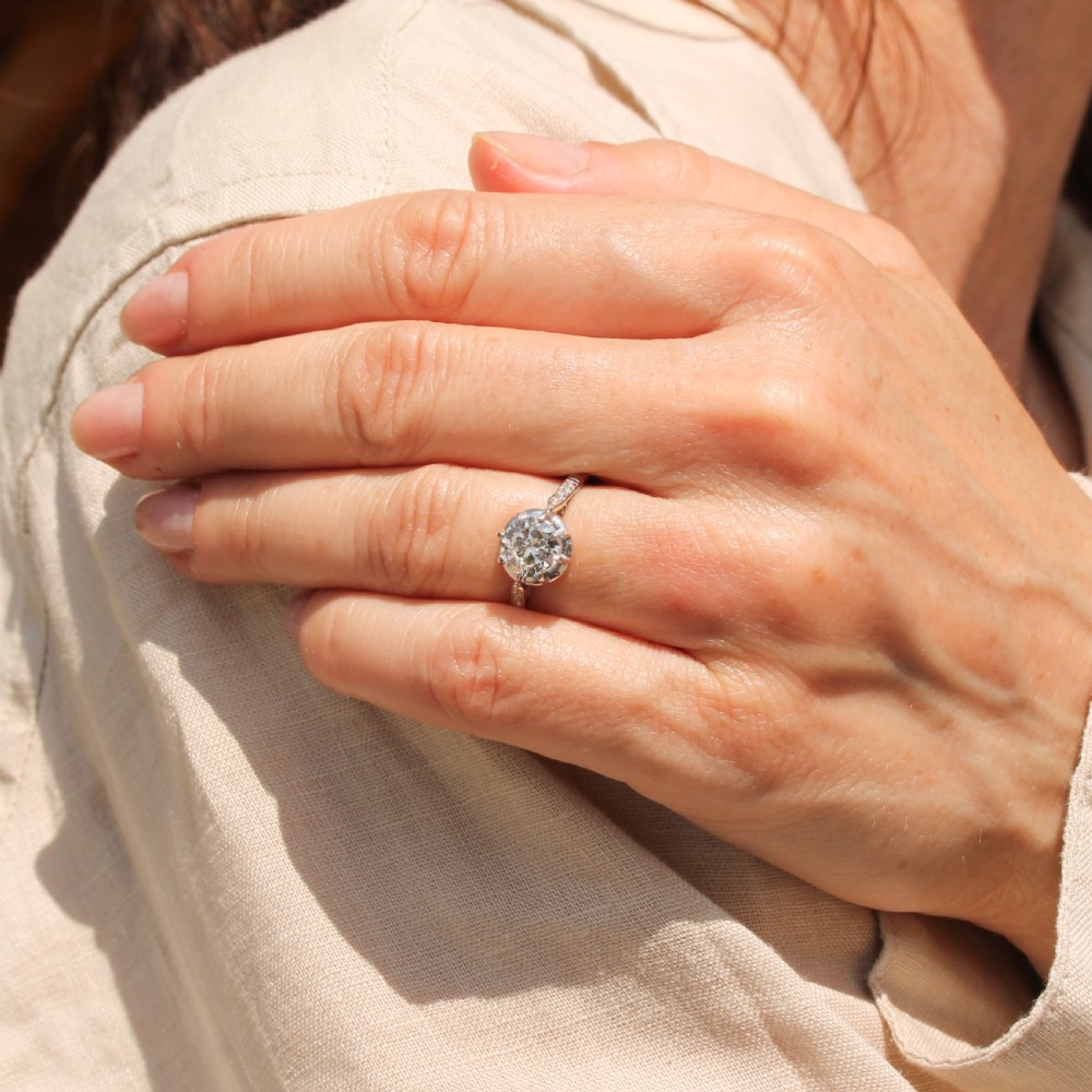 An Art Deco solitaire engagement ring featuring a central round old cut diamond set in white metal with accent diamonds set into the shoulders. View VII