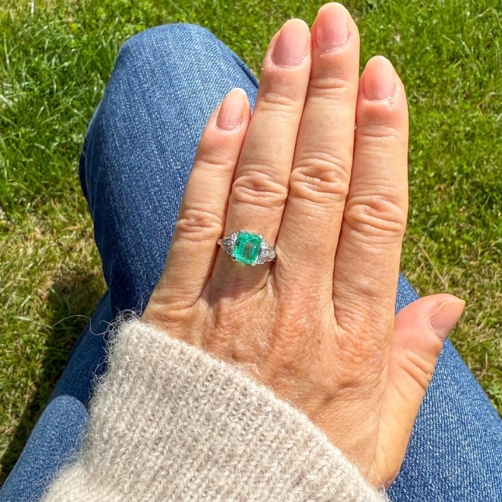 An Art Deco platinum ring featuring a central emerald-cut green gemstone flanked by geometric diamond-set shoulders. View VI