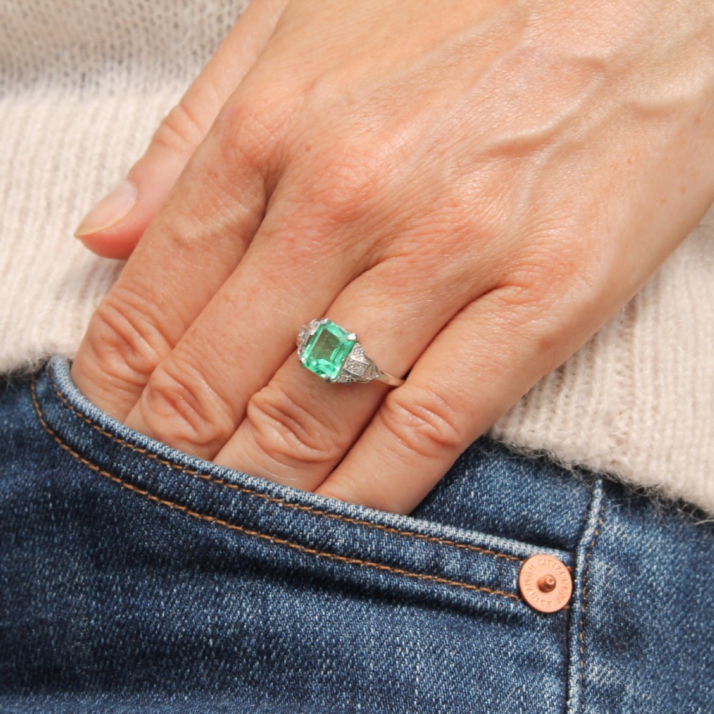 An Art Deco platinum ring featuring a central emerald-cut green gemstone flanked by geometric diamond-set shoulders. View VII
