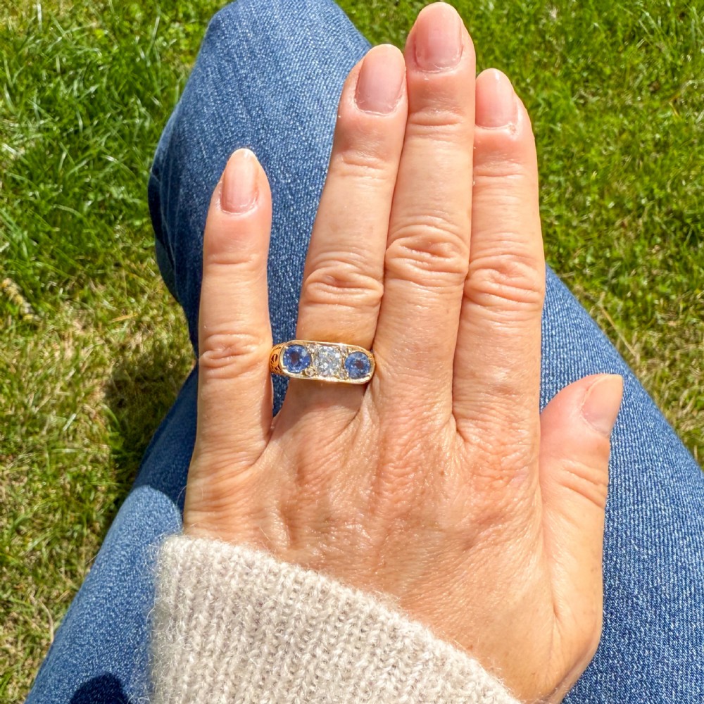 An Art Nouveau yellow gold ring set with a central round old cut diamond flanked by two blue sapphires within an openwork scrollwork gallery. View VII