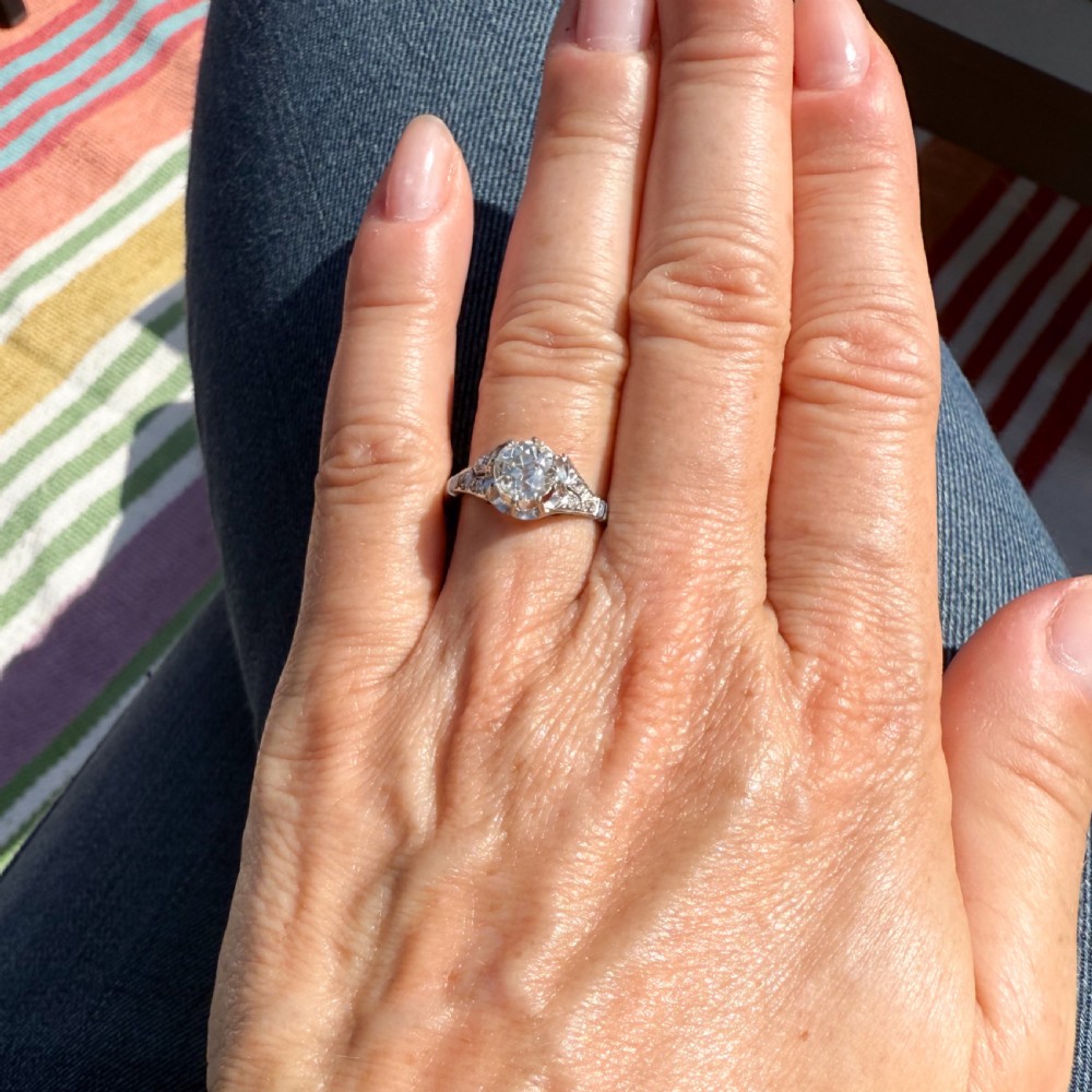 An Art Deco style white metal engagement ring featuring a central round old cut diamond flanked by smaller accent diamonds on sculpted shoulders. View VI