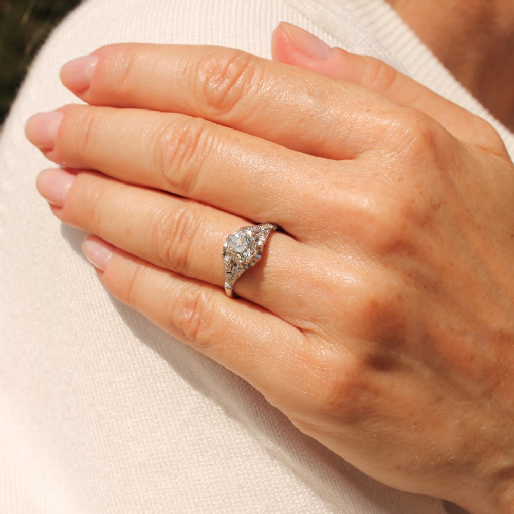 An Art Deco style white metal engagement ring featuring a central round old cut diamond flanked by smaller accent diamonds on sculpted shoulders. View VII