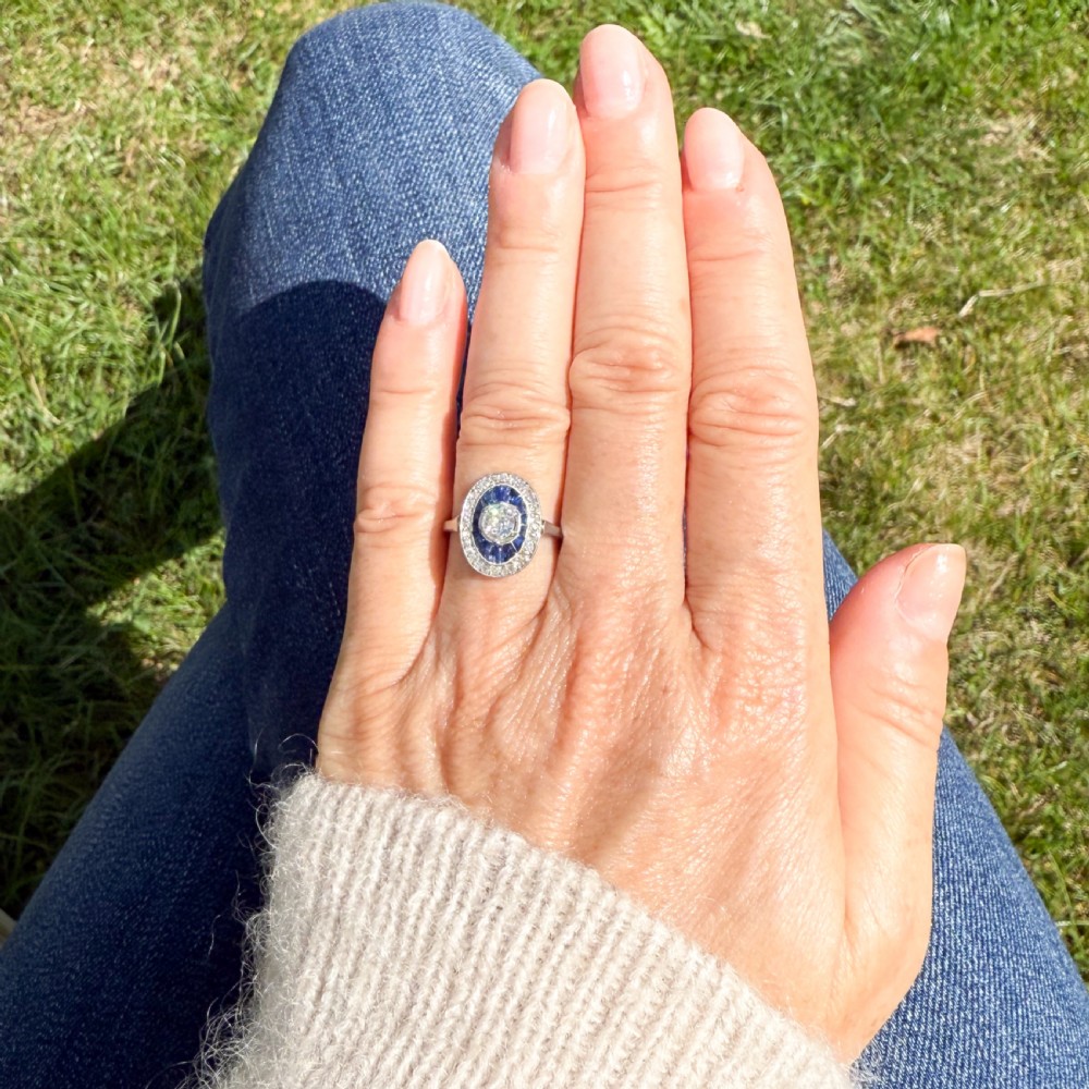 An Art Deco oval cluster ring featuring a central round diamond surrounded by a ring of calibre-cut blue sapphires and an outer diamond halo on a platinum band. View VI