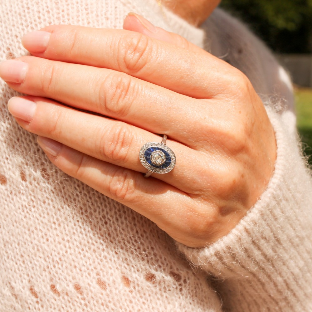 An Art Deco oval cluster ring featuring a central round diamond surrounded by a ring of calibre-cut blue sapphires and an outer diamond halo on a platinum band. View VII
