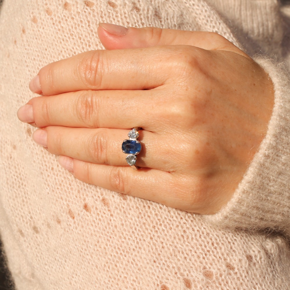 An Art Deco style ring featuring a central oval blue sapphire flanked by two round diamonds set in a silver-toned metal band. View VI