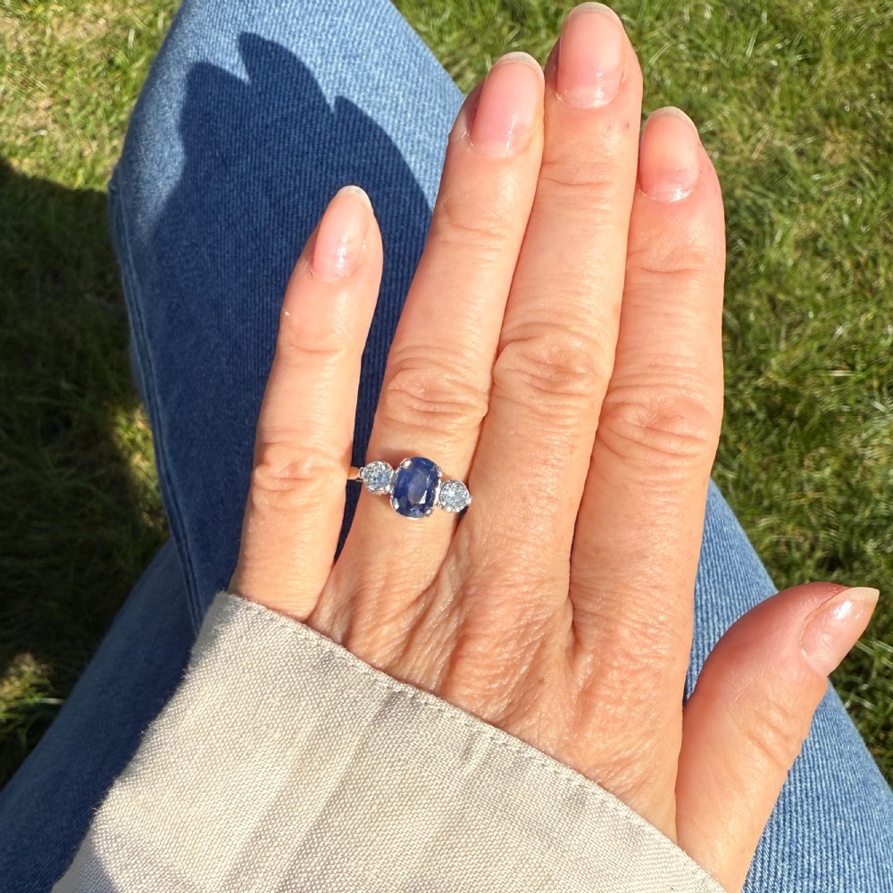 An Art Deco style ring featuring a central oval blue sapphire flanked by two round diamonds set in a silver-toned metal band. View VII
