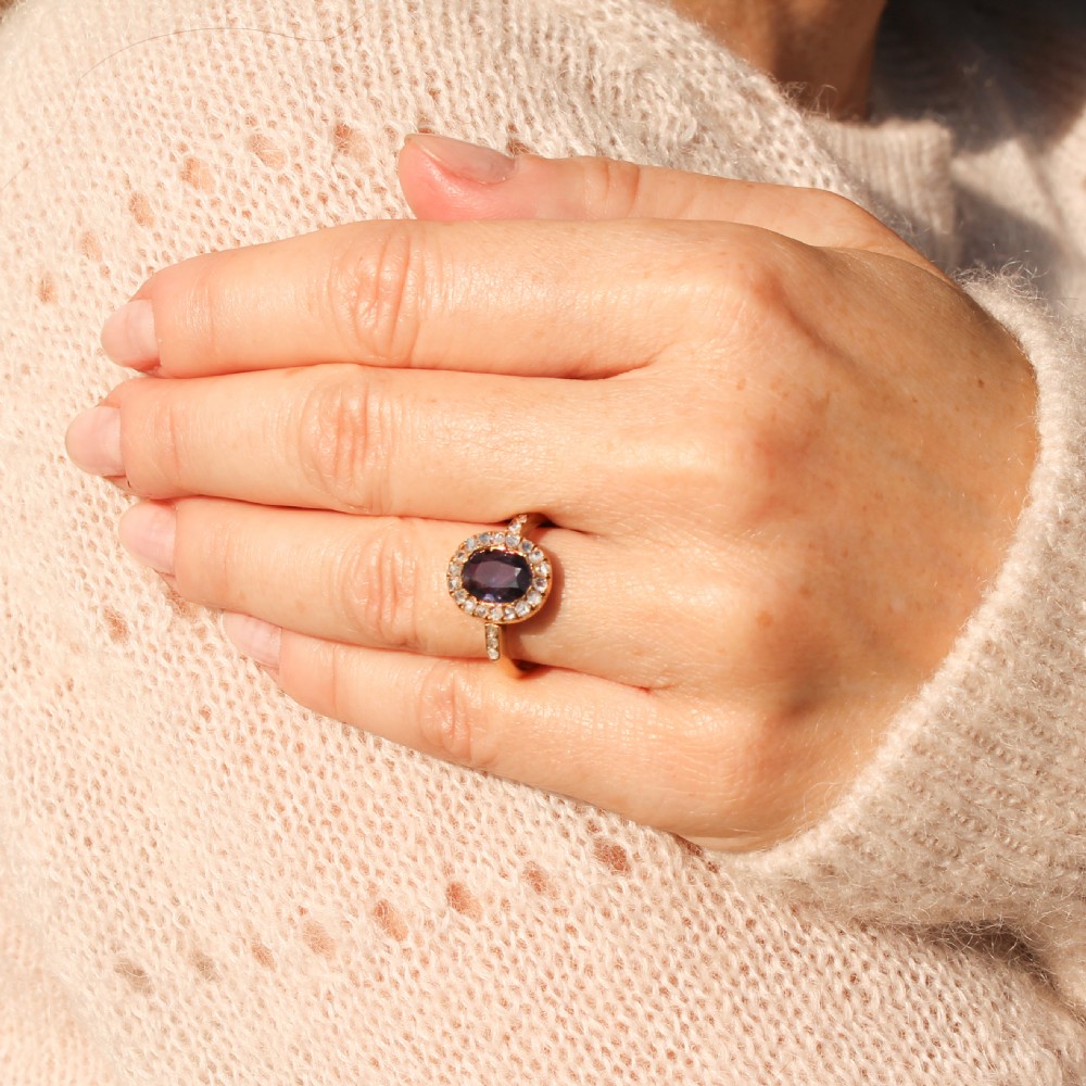 An antique yellow gold ring featuring a central oval blue sapphire surrounded by a halo of small diamonds, with additional diamonds set into the shoulders. View VII