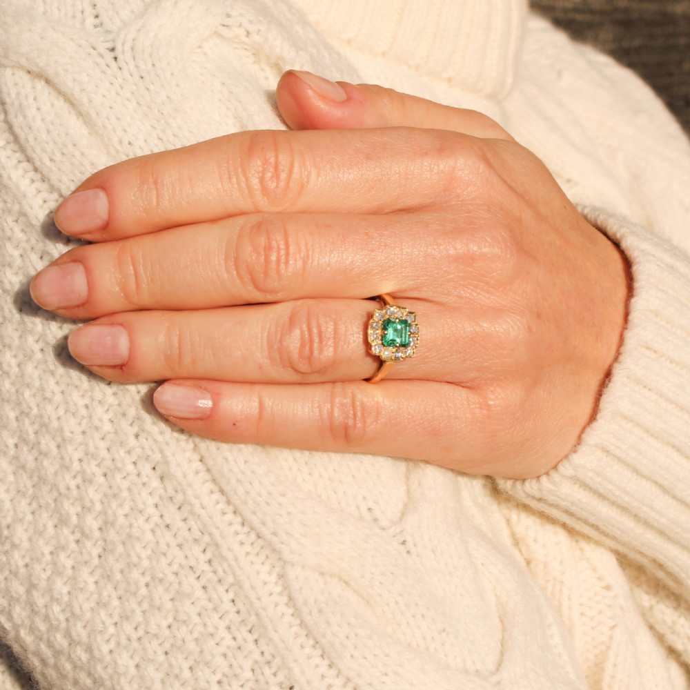 A yellow gold Art Deco cluster ring featuring a central square green emerald surrounded by a geometric halo of round diamonds. View VII