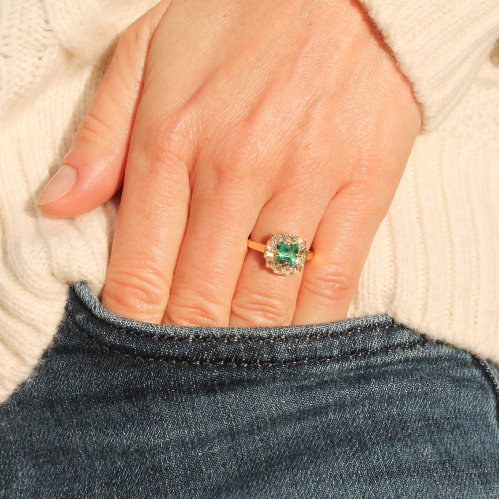 A yellow gold Art Deco cluster ring featuring a central square green emerald surrounded by a geometric halo of round diamonds. View VIII