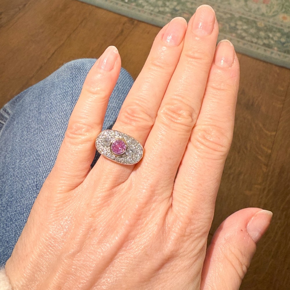 A vintage gold ring featuring a central bezel-set pink sapphire flanked by two round diamonds within a pave-set diamond oval plaque. View II