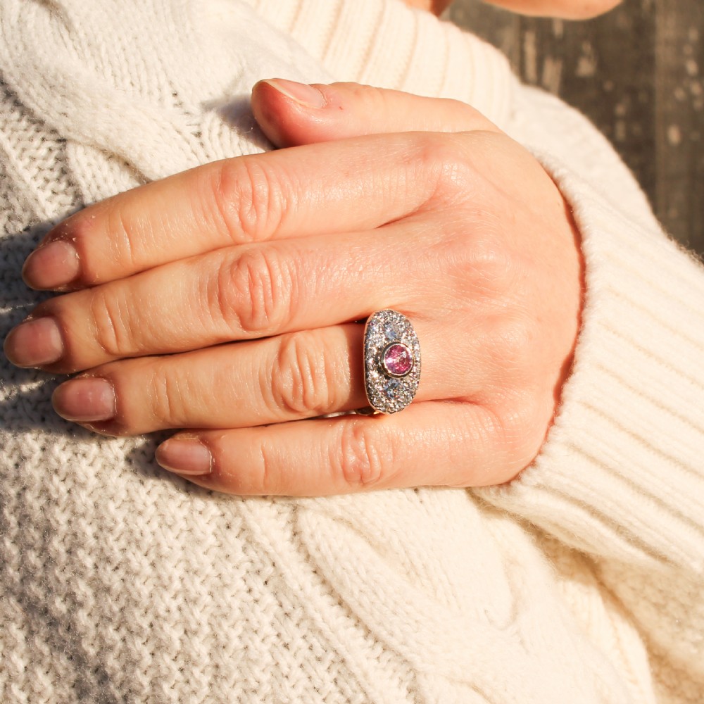 A vintage gold ring featuring a central bezel-set pink sapphire flanked by two round diamonds within a pave-set diamond oval plaque. View VII