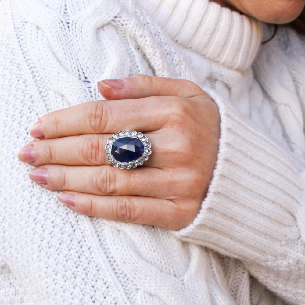 An oval blue sapphire gemstone set in a silver-toned metal ring, surrounded by a halo of circular white diamonds. View VI