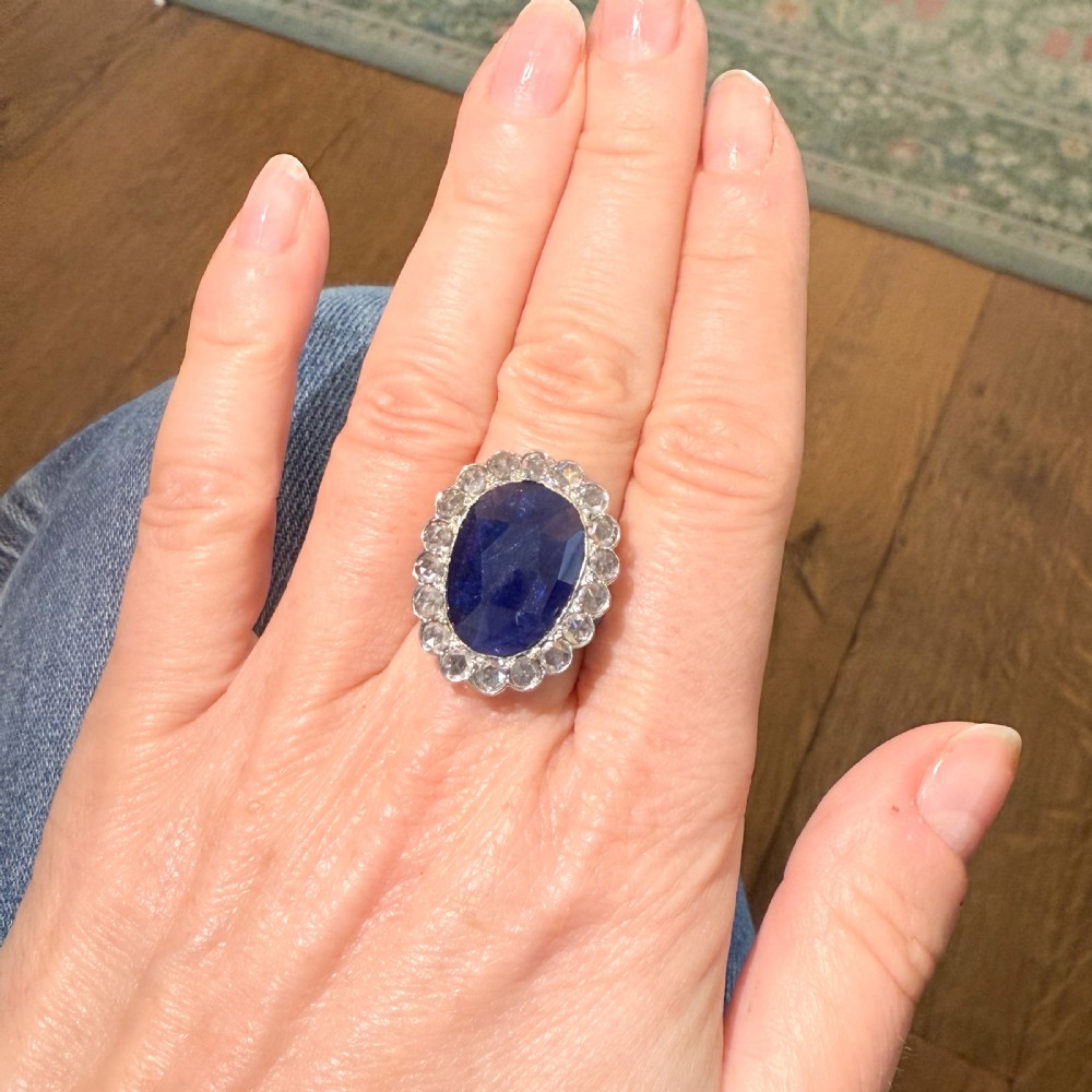 An oval blue sapphire gemstone set in a silver-toned metal ring, surrounded by a halo of circular white diamonds. View VII