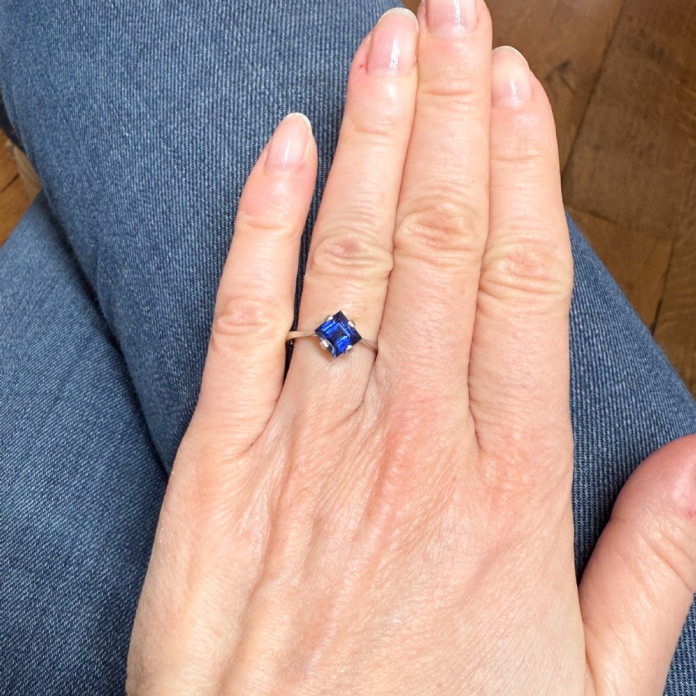A vintage Art Deco platinum solitaire ring featuring a square step-cut blue sapphire mounted on point in claw settings. View VI