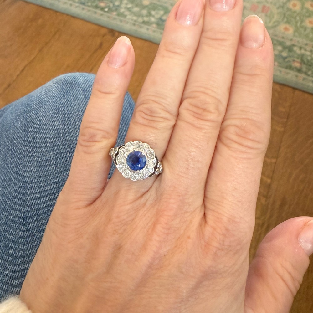 A vintage platinum ring featuring a central round blue sapphire surrounded by a halo of brilliant diamonds with ornate shoulders. View VI