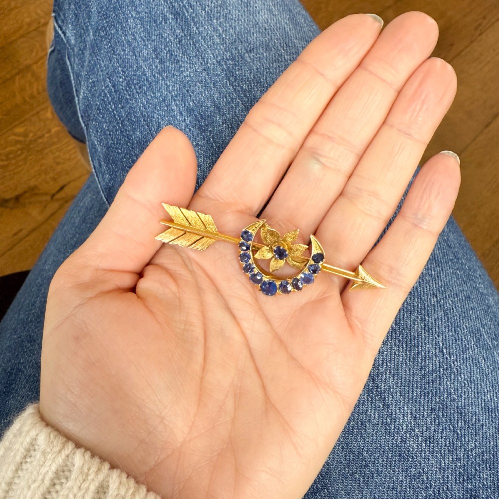 A gold Victorian bar brooch designed as an arrow, featuring a central crescent motif set with blue sapphires around a floral centrepiece. View IX