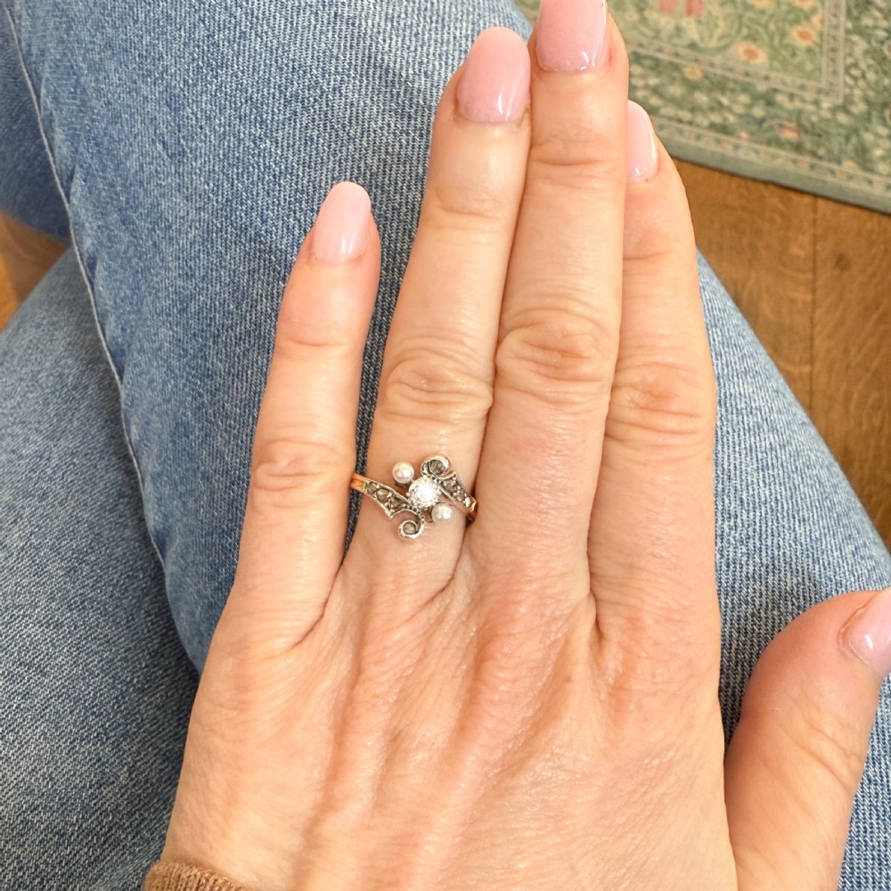 An Art Nouveau gold ring featuring a central old cut diamond surrounded by swirling silver-topped accents set with two pearls and smaller diamonds. View VI