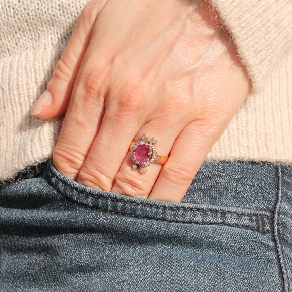 An antique Edwardian ring featuring an oval faceted pink sapphire surrounded by small diamonds set in scrollwork atop a yellow gold band. View II
