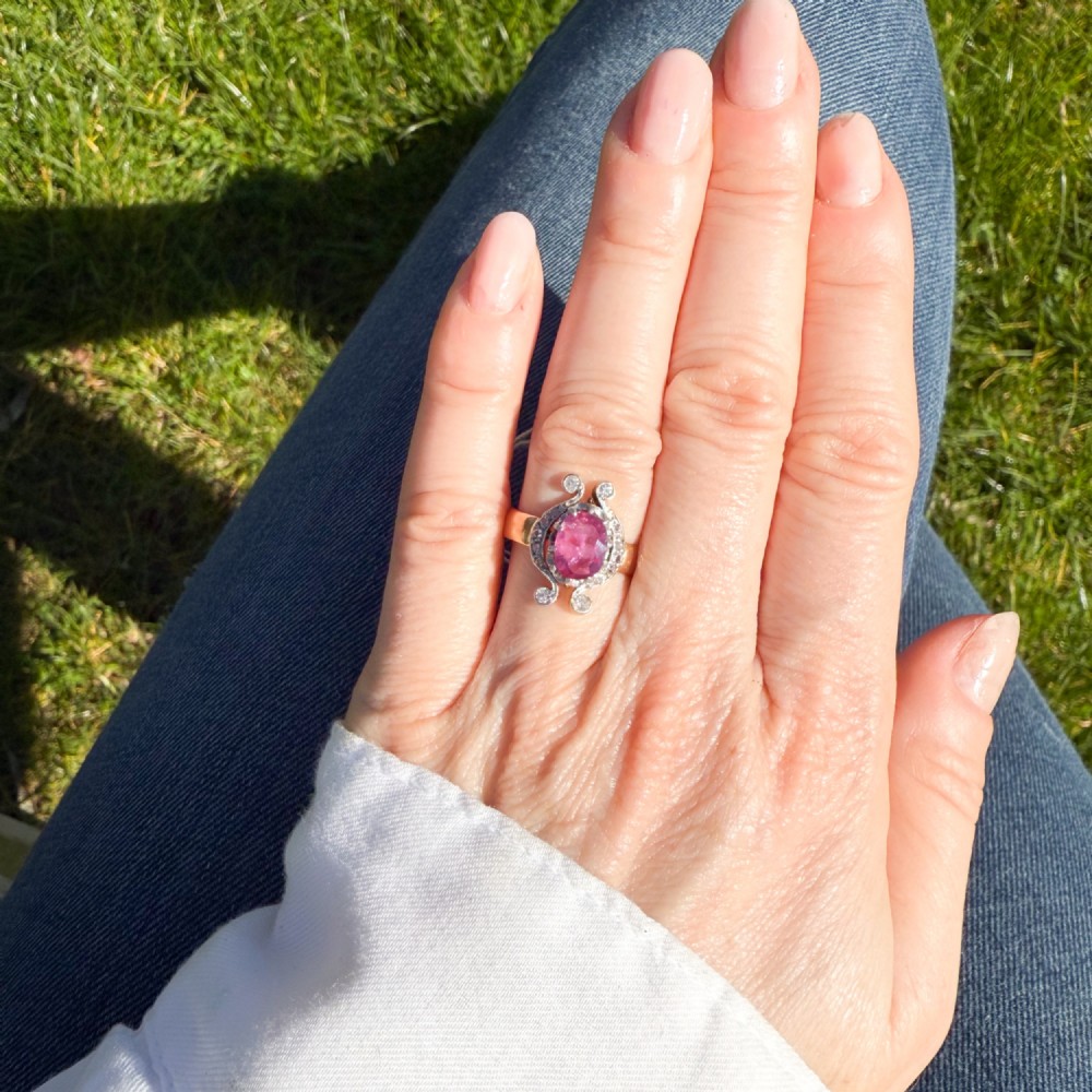 An antique Edwardian ring featuring an oval faceted pink sapphire surrounded by small diamonds set in scrollwork atop a yellow gold band. View VI