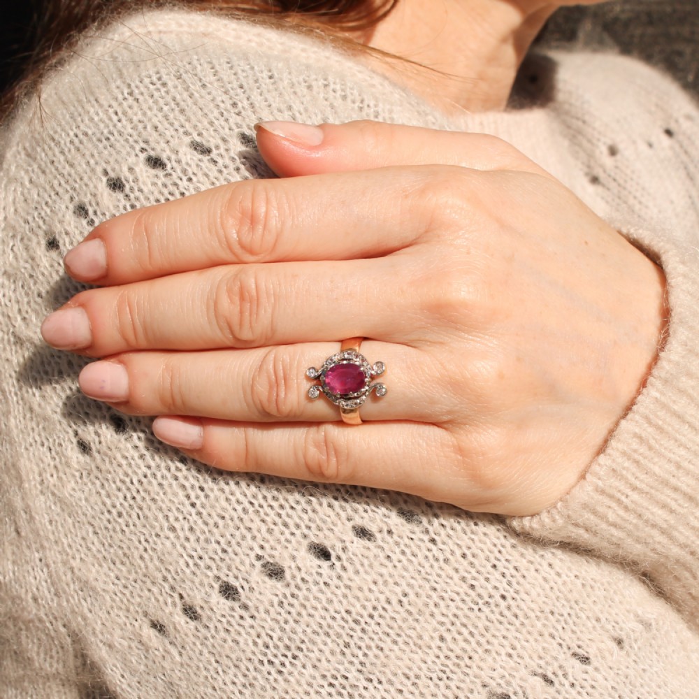An antique Edwardian ring featuring an oval faceted pink sapphire surrounded by small diamonds set in scrollwork atop a yellow gold band. View VII