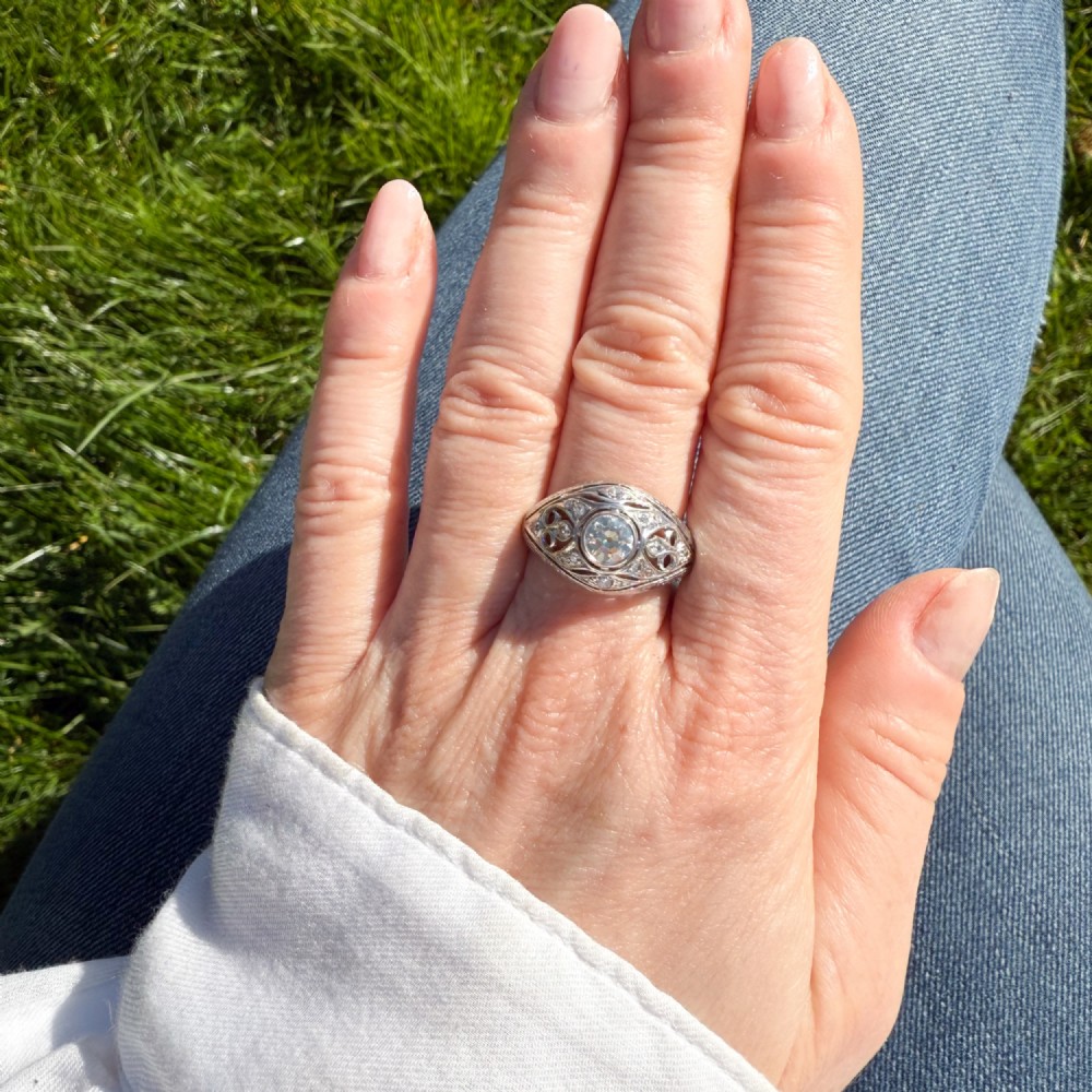 An Art Deco platinum ring featuring a central round old cut diamond in a bezel setting surrounded by pierced filigree scrollwork and small accent diamonds. View VI