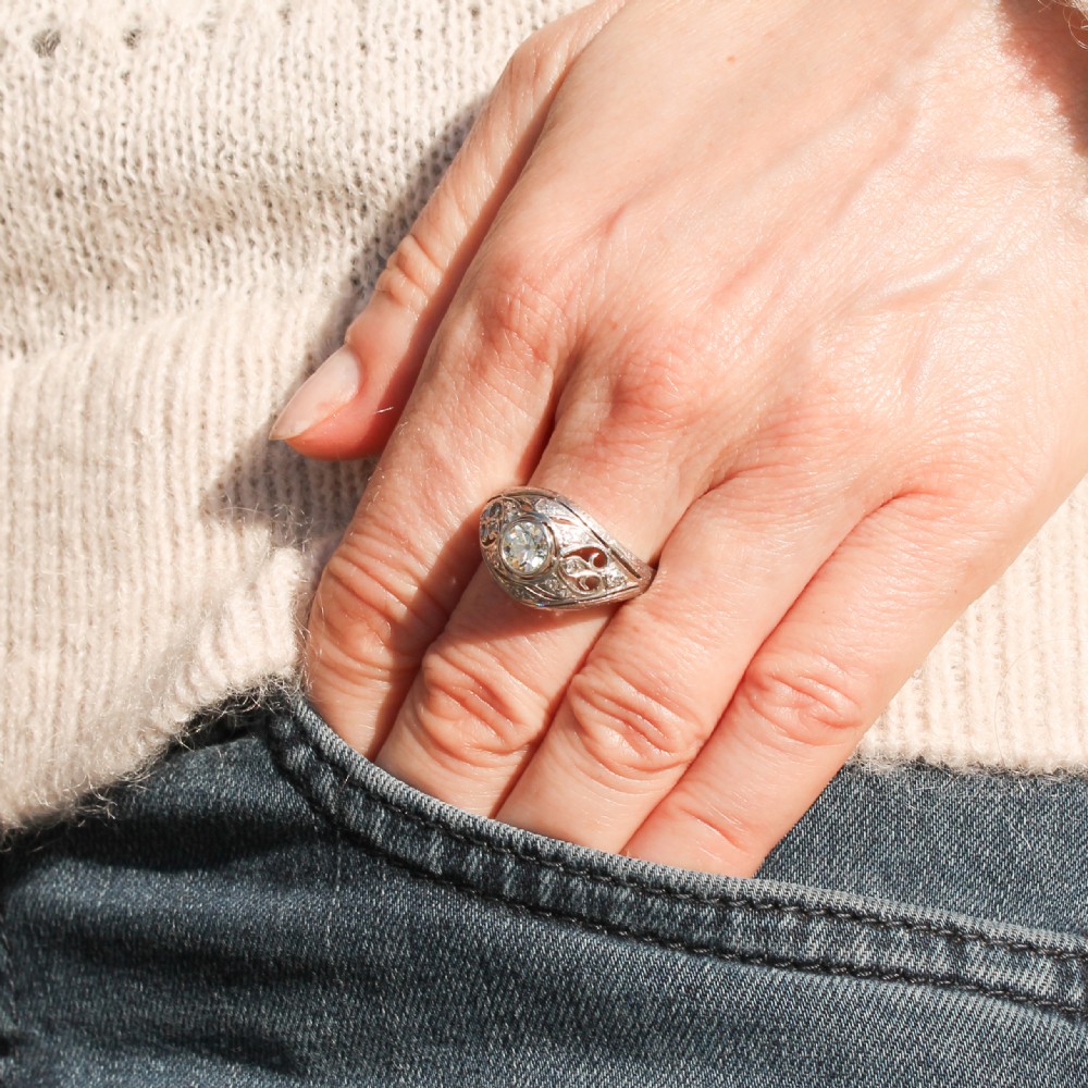An Art Deco platinum ring featuring a central round old cut diamond in a bezel setting surrounded by pierced filigree scrollwork and small accent diamonds. View VII