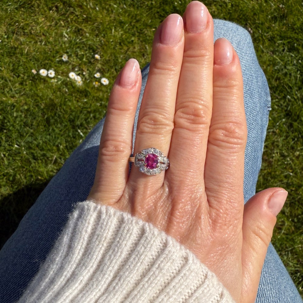 An Art Deco platinum ring featuring an oval pink sapphire in a four-claw setting surrounded by a geometric halo of diamonds with milgrain detail. View VI