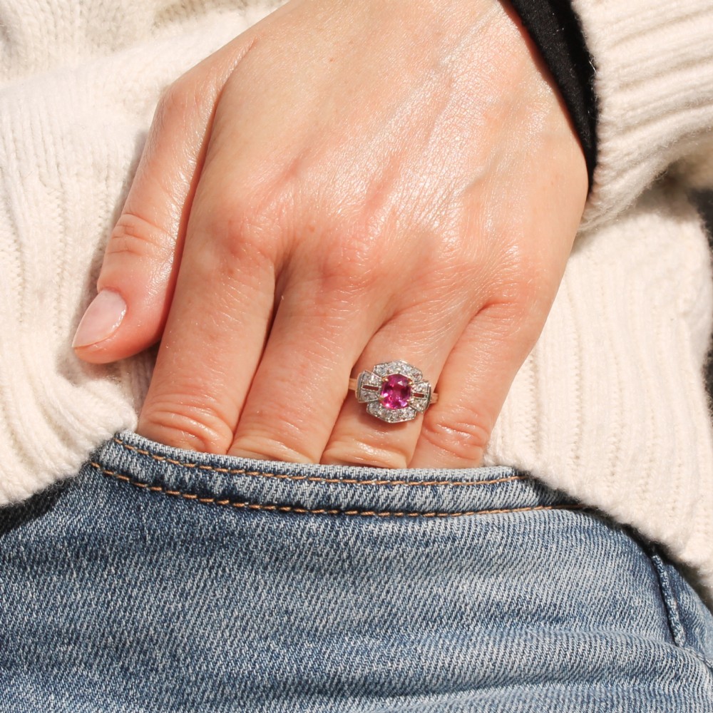 An Art Deco platinum ring featuring an oval pink sapphire in a four-claw setting surrounded by a geometric halo of diamonds with milgrain detail. View VII