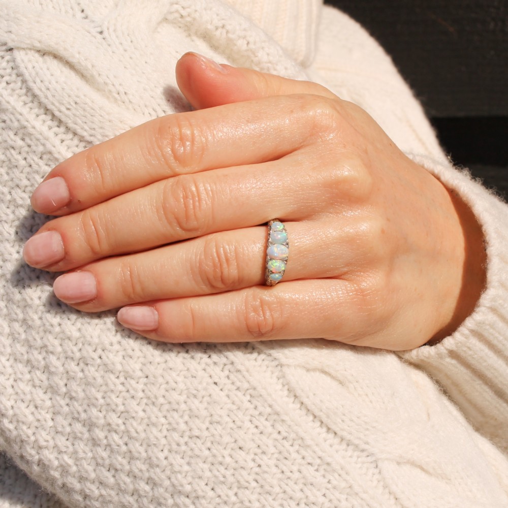 A white metal ring featuring five graduated oval cabochon opals set in a row across a decorative scrollwork setting. View VI