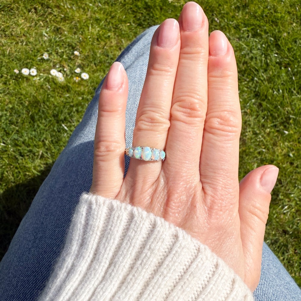 A white metal ring featuring five graduated oval cabochon opals set in a row across a decorative scrollwork setting. View VII
