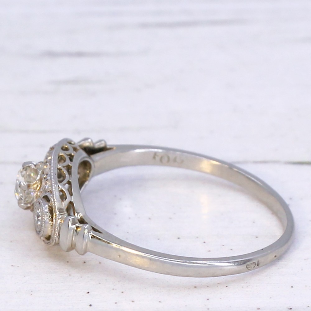 A vintage Art Deco white metal ring featuring a central old cut diamond flanked by two smaller diamonds in an elongated milgrained setting. View IV
