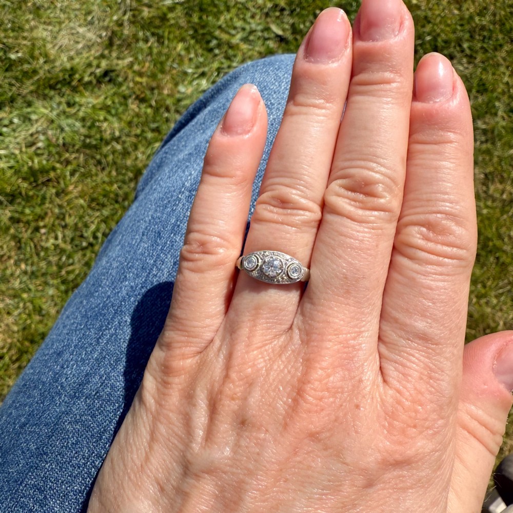 A vintage Art Deco white metal ring featuring a central old cut diamond flanked by two smaller diamonds in an elongated milgrained setting. View VI