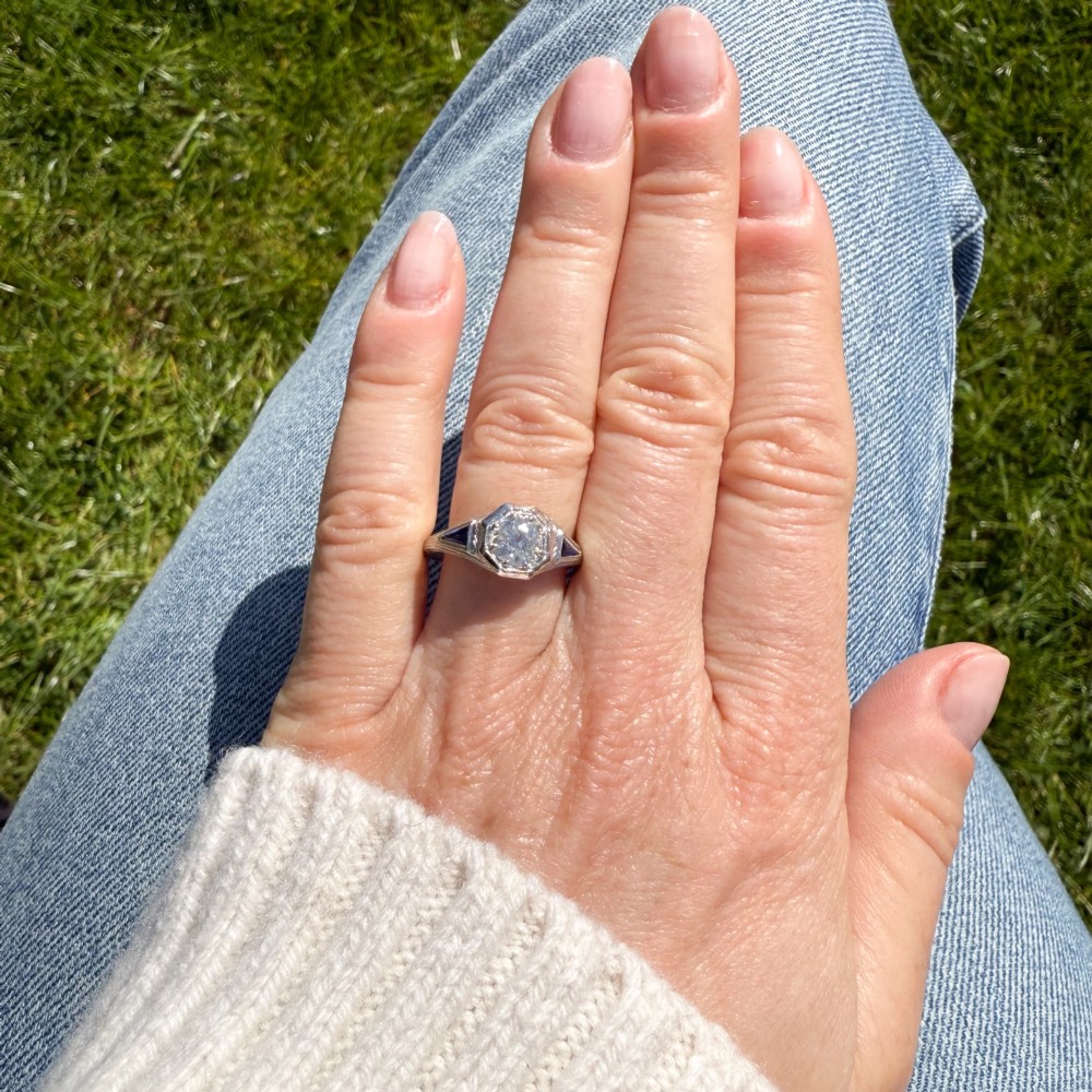 A white metal ring featuring a central old-cut diamond set within an octagonal bezel, flanked by triangular blue sapphires and fine engraved detailing along the band. View VI