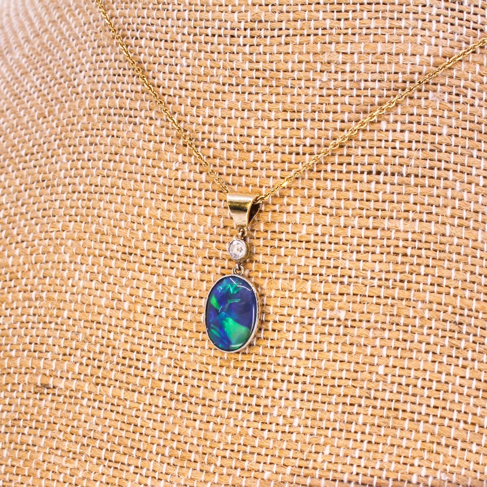 An Edwardian black opal and diamond pendant featuring an oval cabochon opal with blue and green play-of-colour mounted in gold below a bezel-set diamond.