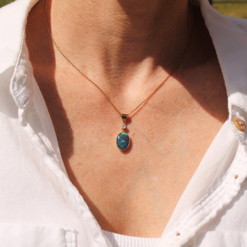An Edwardian black opal and diamond pendant featuring an oval cabochon opal with blue and green play-of-colour mounted in gold below a bezel-set diamond. View VI