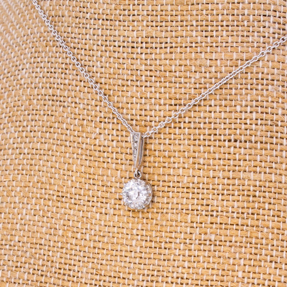 An Art Deco diamond pendant featuring a circular old cut diamond suspended from a linear diamond-set bale on a fine silver-toned chain.