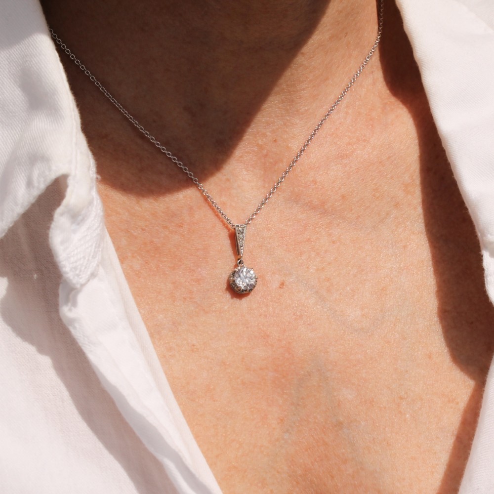 An Art Deco diamond pendant featuring a circular old cut diamond suspended from a linear diamond-set bale on a fine silver-toned chain. View II