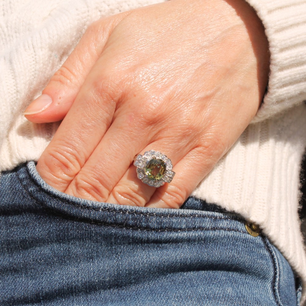 A vintage white precious metal ring featuring a central oval green sapphire encircled by a halo of round diamonds and diamond-set shoulders. View II