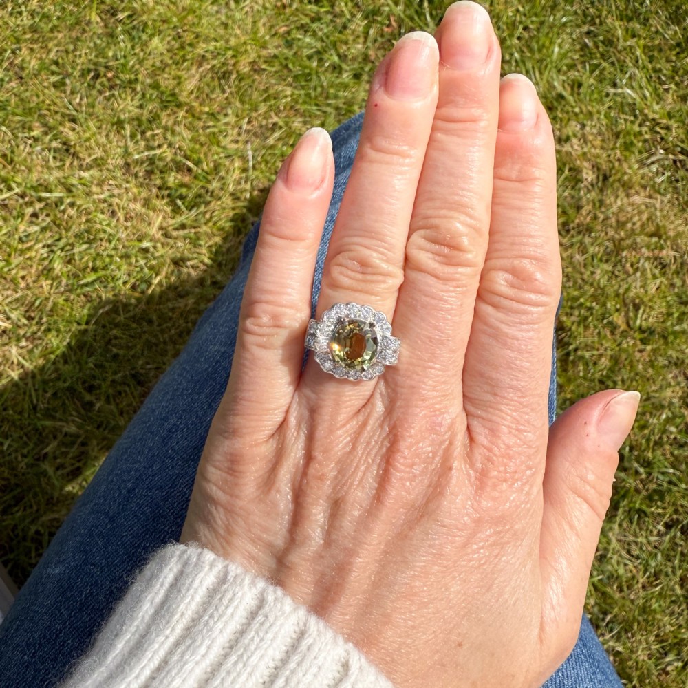 A vintage white precious metal ring featuring a central oval green sapphire encircled by a halo of round diamonds and diamond-set shoulders. View VI