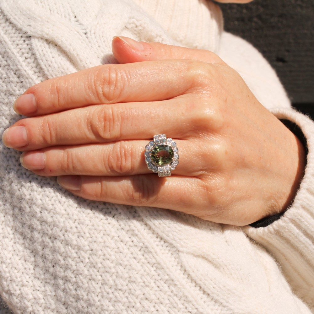 A vintage white precious metal ring featuring a central oval green sapphire encircled by a halo of round diamonds and diamond-set shoulders. View VII