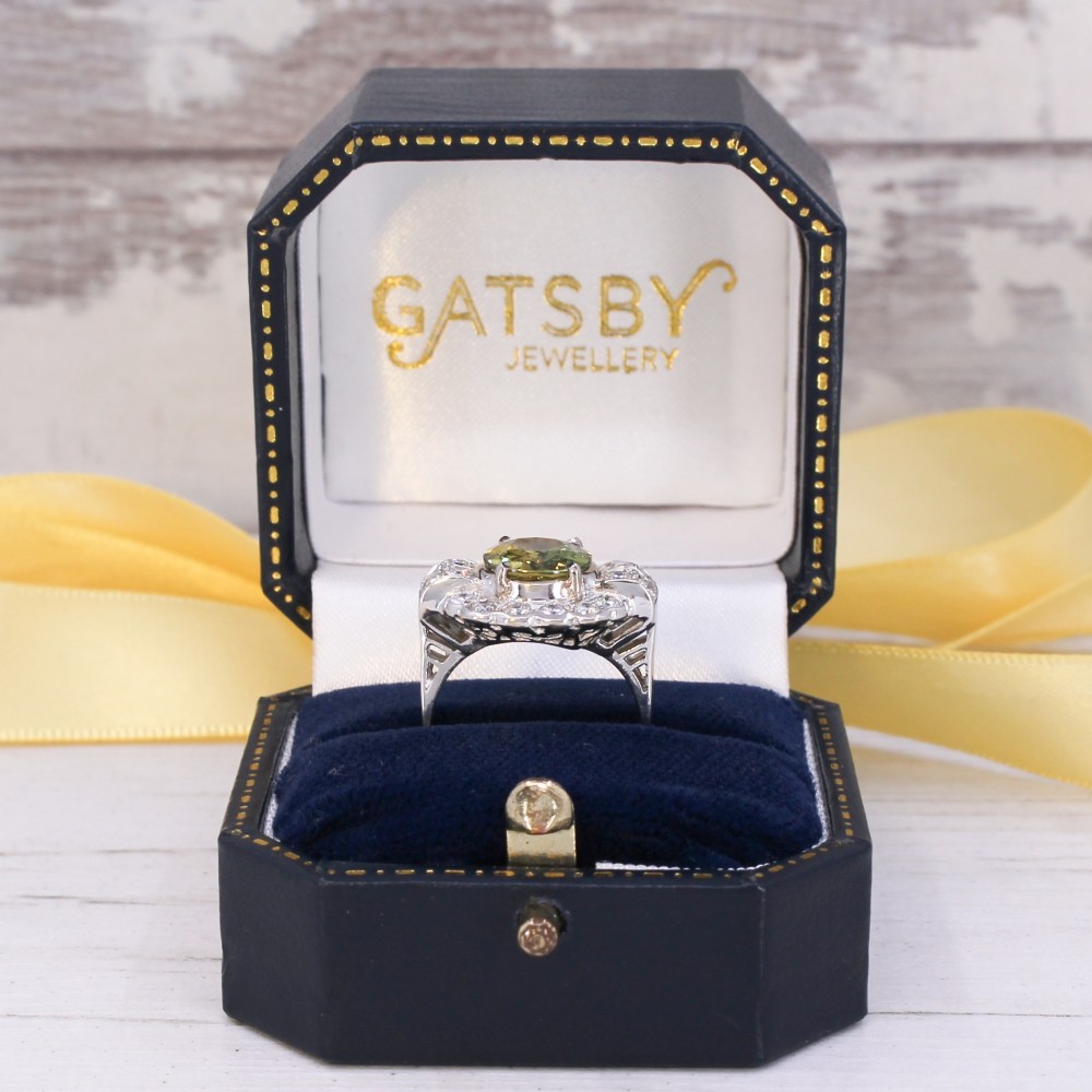 A vintage white precious metal ring featuring a central oval green sapphire encircled by a halo of round diamonds and diamond-set shoulders. View IX