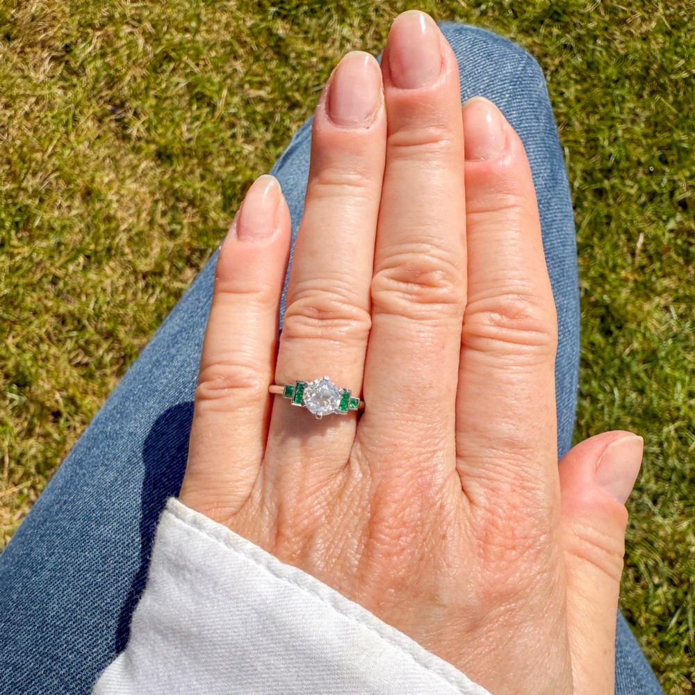 A platinum ring set with a central round old European cut diamond flanked by channel-set green emerald shoulders. View VI