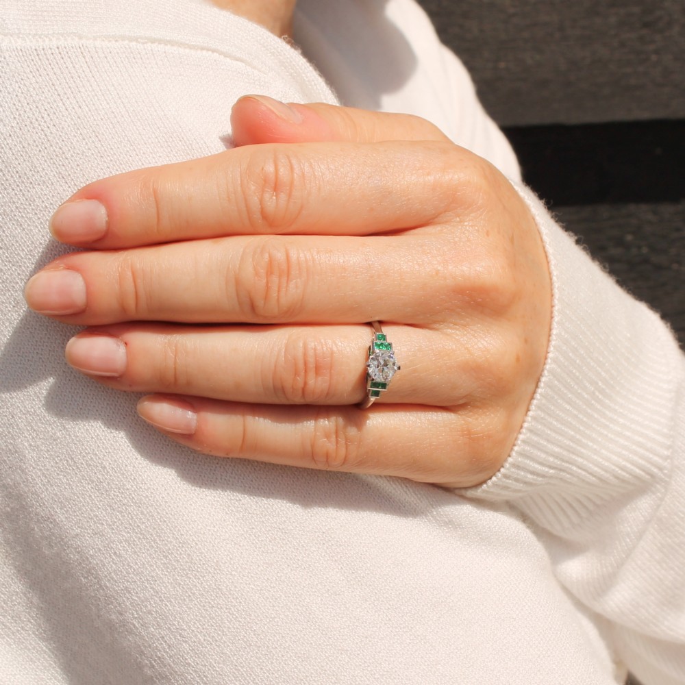 A platinum ring set with a central round old European cut diamond flanked by channel-set green emerald shoulders. View VII