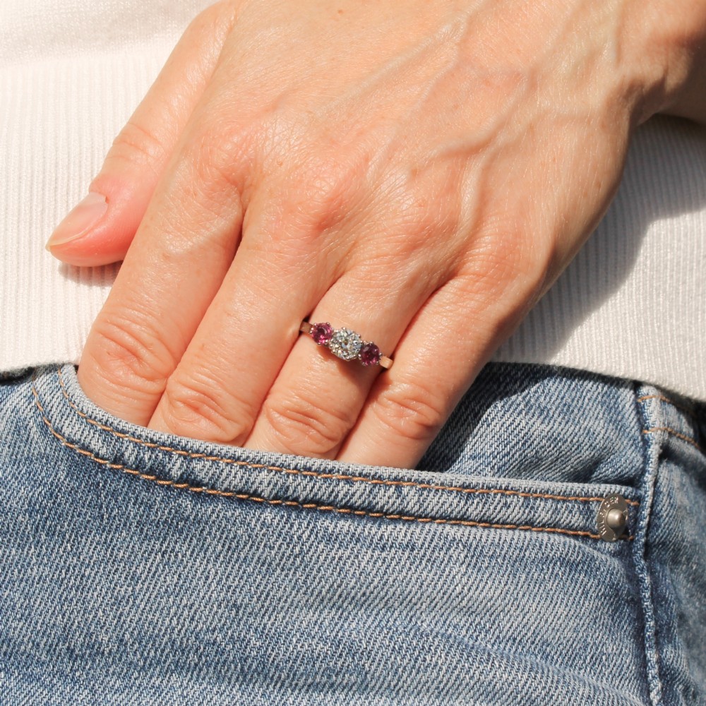 A white precious metal three-stone ring featuring a central round diamond flanked by two red rubies. View II