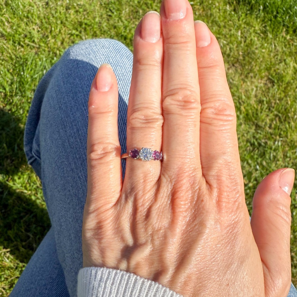 A white precious metal three-stone ring featuring a central round diamond flanked by two red rubies. View VI