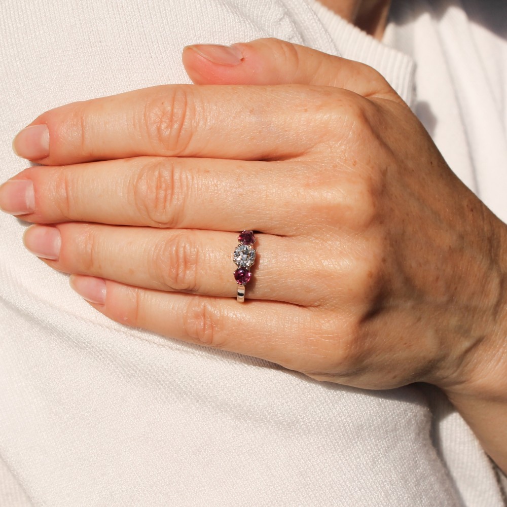 A white precious metal three-stone ring featuring a central round diamond flanked by two red rubies. View VII