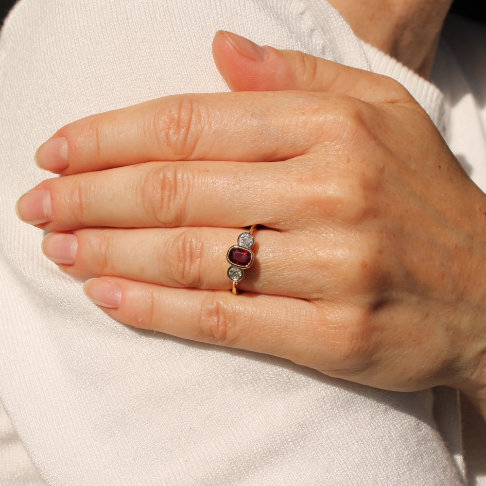 A yellow gold three-stone ring featuring a central cushion cut red ruby set in a bezel, flanked by two old-cut round diamonds. View II