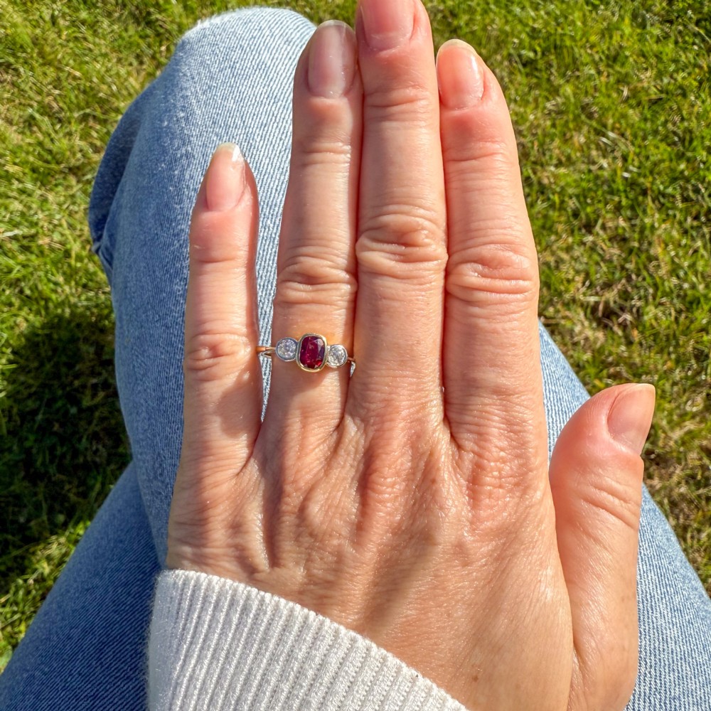 A yellow gold three-stone ring featuring a central cushion cut red ruby set in a bezel, flanked by two old-cut round diamonds. View VI