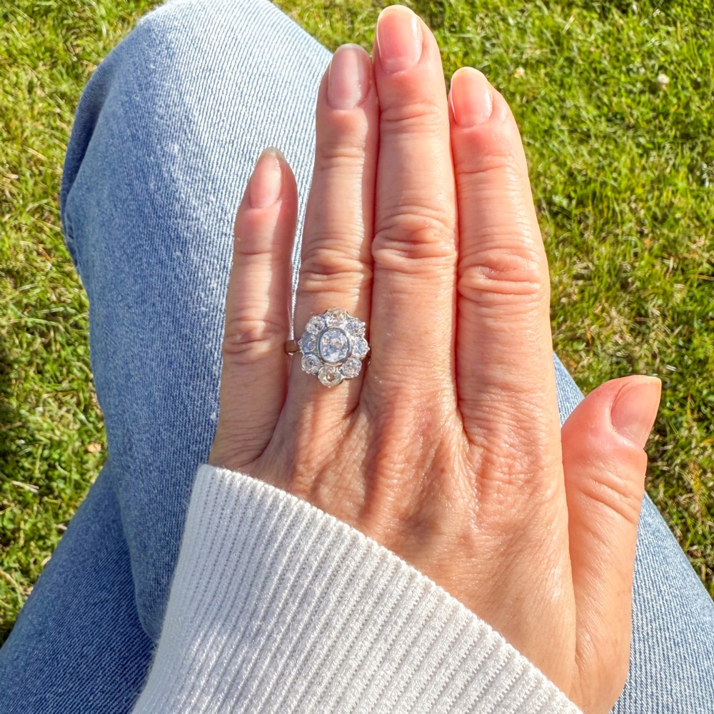 A vintage platinum ring featuring a central old cut diamond surrounded by a halo of eight smaller diamonds in a floral cluster setting on a smooth band. View VI